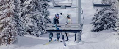 A pair of skiers rides a chairlift while smiling at the camera. 