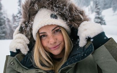 A woman stands in the snow wearing fluffy gloves and a jacket with a faux fur trim smiling at the camera. 