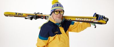 Hotdog Hans is pictured wearing a vintage Columbia Sportswear jacket as he holds a pair of skis and looks at the camera. 