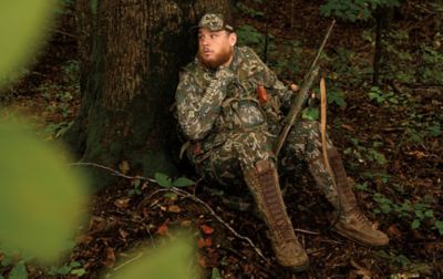 Luke Combs, tests the padded back panel of the Luke Combs Turkey Hunting vest as he leans against a tree, shotgun in hand, scanning for wild turkeys.