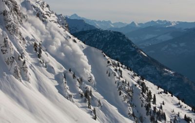 Endless mountain peaks stretch across the horizon, while an avalanche charges down a snow-covered slope, carving its way through the quiet landscape.