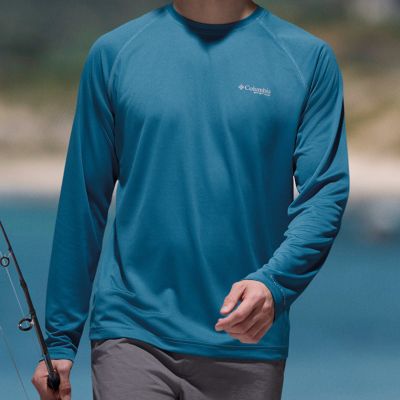 Man walking in a sunny scene wearing a blue long-sleeve shirt and holding a fishing rod.