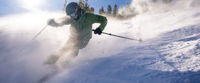 A skier in Columbia Sportswear gear kicks up a cloud of powder while moving across a slope from the sun into shade.