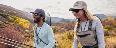 A woman and a man, both wearing protective long sleeve sun shirts and  brimmed ball caps, smile as they carry fly fishing equipment.