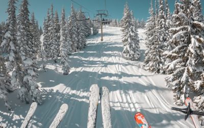 Three skiers are riding up a chairlift. Between their ski tips you can see a snow-laden slope with deep tracks. 