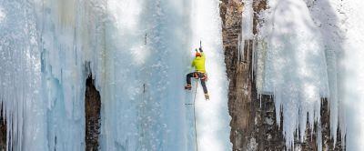 A person wearing crampons and using ice axes climbs a frozen waterfall, secured by a rope and ice screws for protection. 