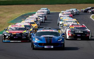 NASCAR race cars form two lines on the track behind a lead car. 

 