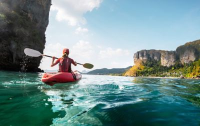 A woman in a red kayak paddles across sparkling blue water. There are steep cliffs of both sides.