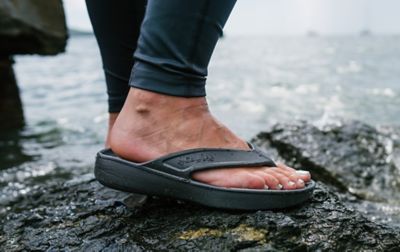 A person with polished toenails wears Columbia flip-flops while standing on a rocky, wet surface.