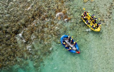 Two whitewater rafts, one yellow and the other blue, each carrying seven people, float side by side down a shallow stretch of river.