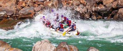 Eight people and their guide, all wearing helmets, paddle in a whitewater raft as they head toward a big wave.