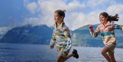 Two kids in colorful fleeces and shorts running by a lake.