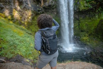 A backpacker pauses to look at a gorgeous cascading waterfall in the middle of a lush green forest. 