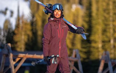 A person in a maroon Columbia Sportswear ski jacket and ski pants walks with skis over their shoulder and ski poles in hand. 