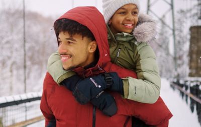 A man in an red jacket carries a child on their back who is wearing Columbia Sportswear mittens. 