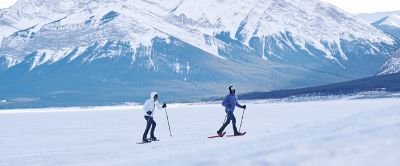 Two people, wearing Columbia Sportswear insulated jackets, snowshoeing across a snowy clearing with large mountains in the background. 