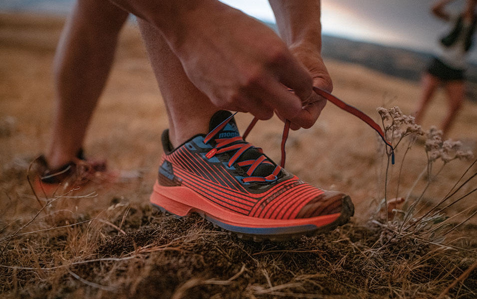 A close-up photo of a trail runner’s hands tying a red-and-blue trail running shoe. 