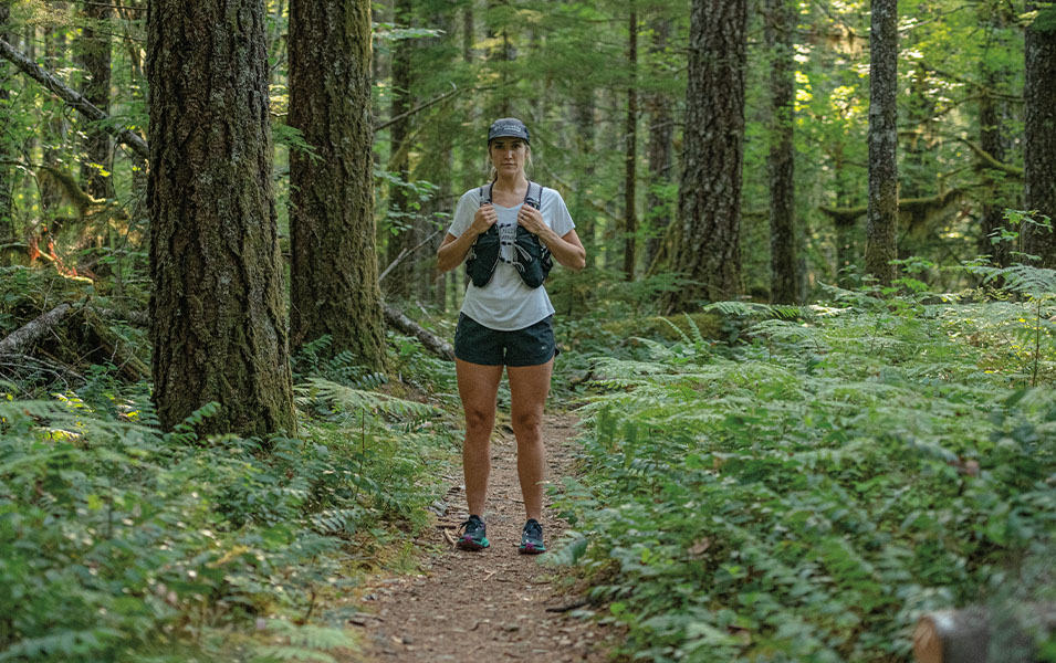 A woman in trail running gear stands in a forest staring at the camera with a bold stance. 