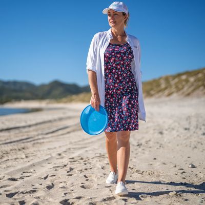 A woman walking on the beach with a frisbee. She's wearing a loose button-down shirt with the Freezer III Dress in floral print.