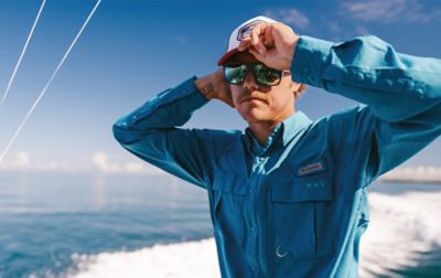 Standing on a boat with wake trailing, a man in a Columbia fishing shirt and sunglasses tugs his ball cap low over his forehead.