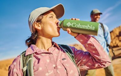 While hiking in the desert, a woman in a patterned Columbia fishing shirt and ballcap sips from her Columbia water bottle.