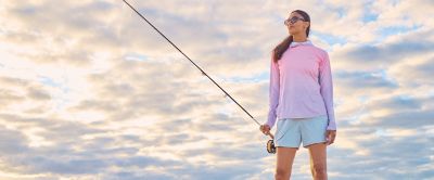 A woman sporting sunglasses, shorts, and a sun-protective hoodie holds a fly rod; her apparel’s colors harmonize with the blue sky and clouds.