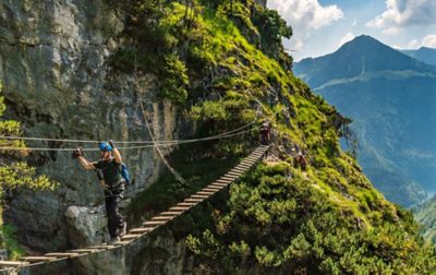 A person walking across a swinging wood and cable suspension bridge high above the ground in rocky, mountainous terrain. The bridge is a part of a via ferrata system and the person is safely attached to the cable so there’s no fear of falling.