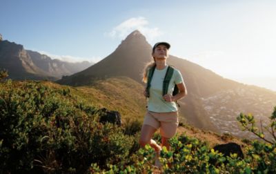 A female hiker with a backpack and sun-protective hat walks along a ridgeline after summiting a nearby peak. 