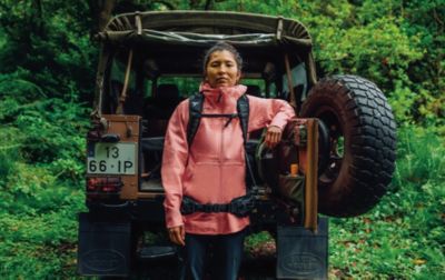 A woman, wearing a pack and a warm jacket,  leans against the back of a 4-wheel drive vehicle parked in a forest.