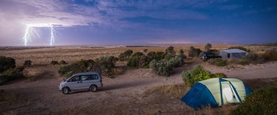 A camping van is parked next to a tent that’s pitched along a dirt road. Above a planted field, the sky has turned a dark shade of blue and a thundercloud hovers above two spears of lightning that are extending to the ground.