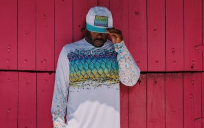 A man leans against a wooden wall, wearing a ball cap and sun-protective hoodie with distinctive Bentley graphics.