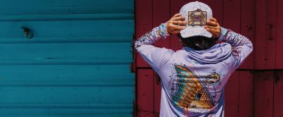 A person wears a lavender sun-protective hoodie and matching ball cap, both featuring marine life graphics. 