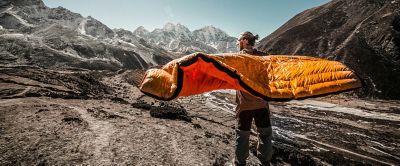 Person standing in front of big mountains, with a sleeping bag draped over his shoulders and arms. The wind is blowing the sleeping bag and helping to air and dry it out. 
