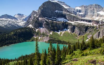 A pristine forest surrounds a round glacial lake at the base of a snow-covered granite peak.
