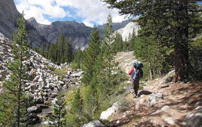 A hiker with a large pack and trekking poles walks alongside a rocky creek toward large granite peaks.