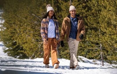 man an woman wearing Columbia Hiking pants walk down a snowy covered path. 