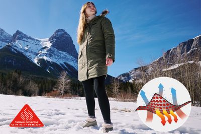A woman stands in front of a snowy landscape with mountains. She's staying warm in a thermal-reflective jacket.