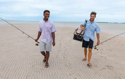 Two men in shorts and sandals are carrying fishing poles as they walk along a sandy beach.