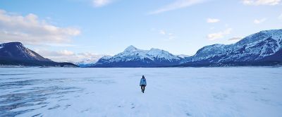 A lone person stands in a vast snow covered valley. Behind the person is a snow-covered mountain range with large, jagged peaks. 