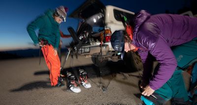 Blurry mid motion image of three skiers getting ready at the back of their truck, putting on ski boots and bibs at dusk.