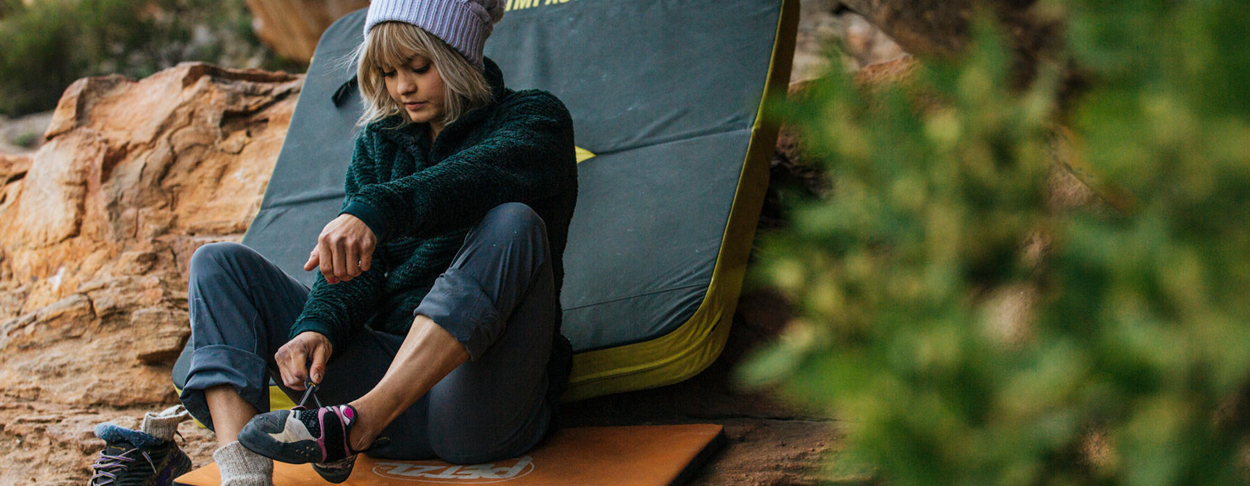 A woman putting on her climbing shoes, sitting on a crash pad, in the Women's Women's Monkey Fleece™ Pullover. 
